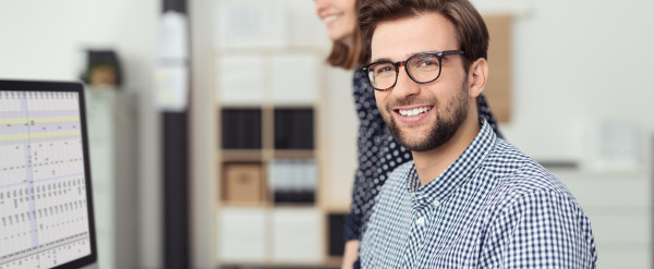 Kaufmännischer Mitarbeiter arbeitet am Computer mit Tabellenansicht, während eine Kollegin im modernen Büro unterstützt.