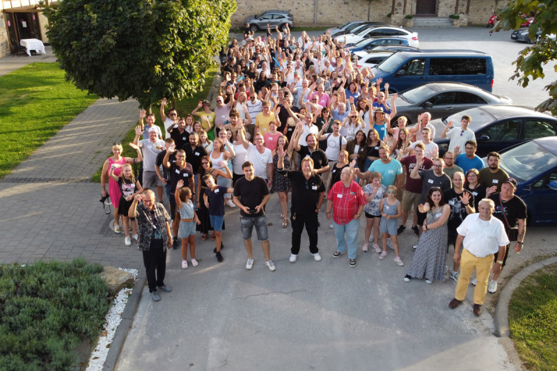 Große Gruppe von Mitarbeitenden und Familienmitgliedern steht im Freien zusammen und winkt in die Kamera bei einem sommerlichen Firmenevent.