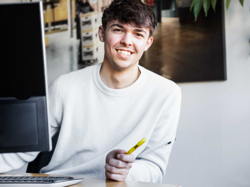 Person arbeitet am Schreibtisch vor einem Computer und hält einen gelben Marker in der Hand.