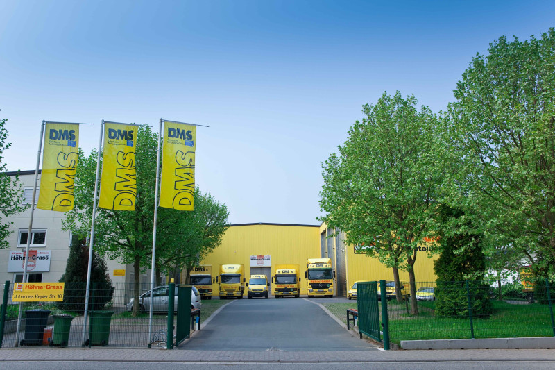 Standort der Höhne‑Grass GmbH mit DMS‑Flaggen und gelben Umzugsfahrzeugen.