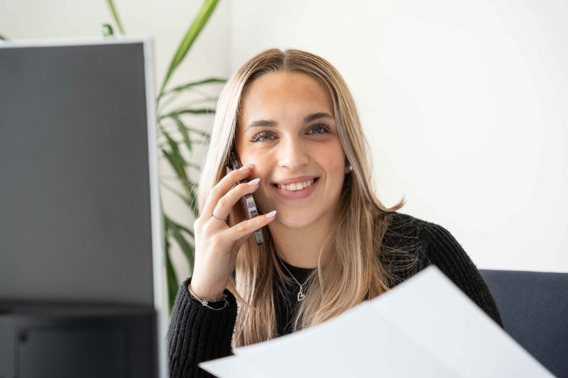 Auszubildende telefoniert am Schreibtisch und arbeitet mit Unterlagen am Computer.
