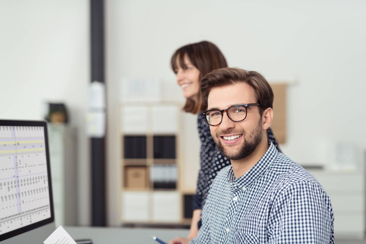 Mitarbeiter sitzt an einem Schreibtisch in einem hellen Büro und schaut in die Kamera.