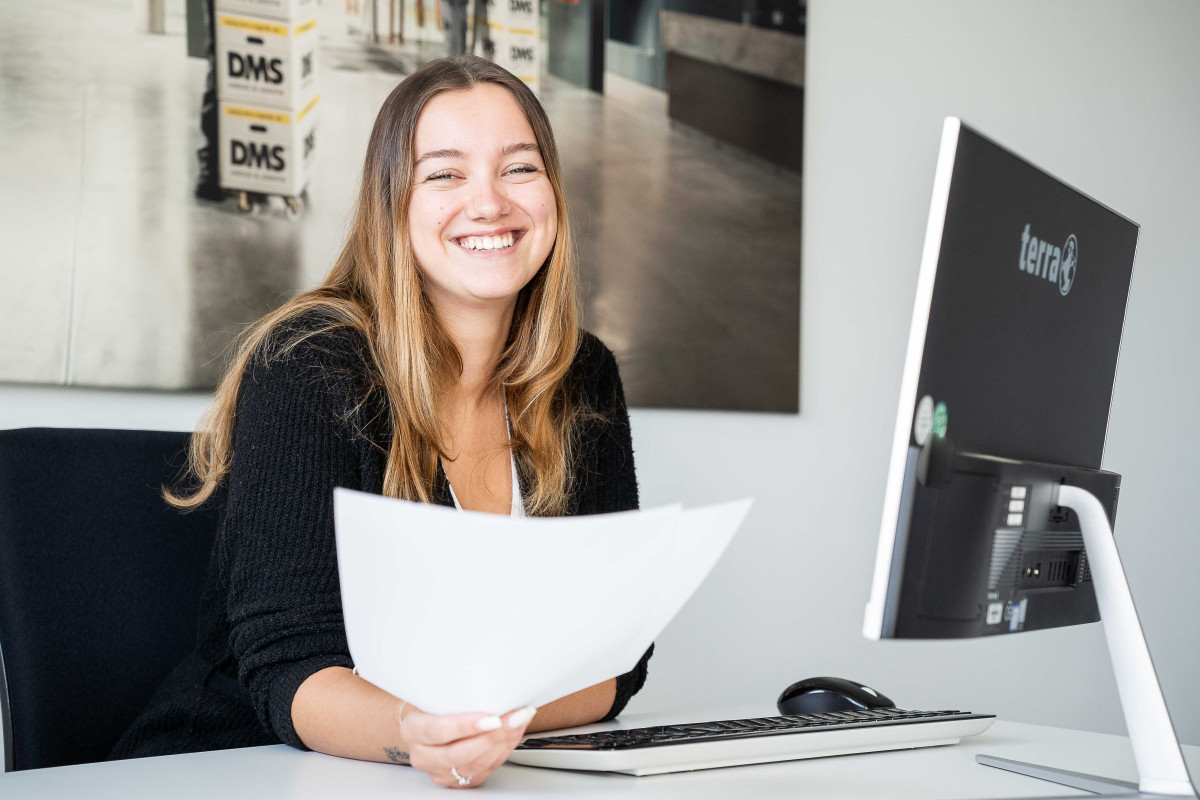 Mitarbeiterin sitzt am Schreibtisch, arbeitet am Computer und hält Unterlagen in der Hand.