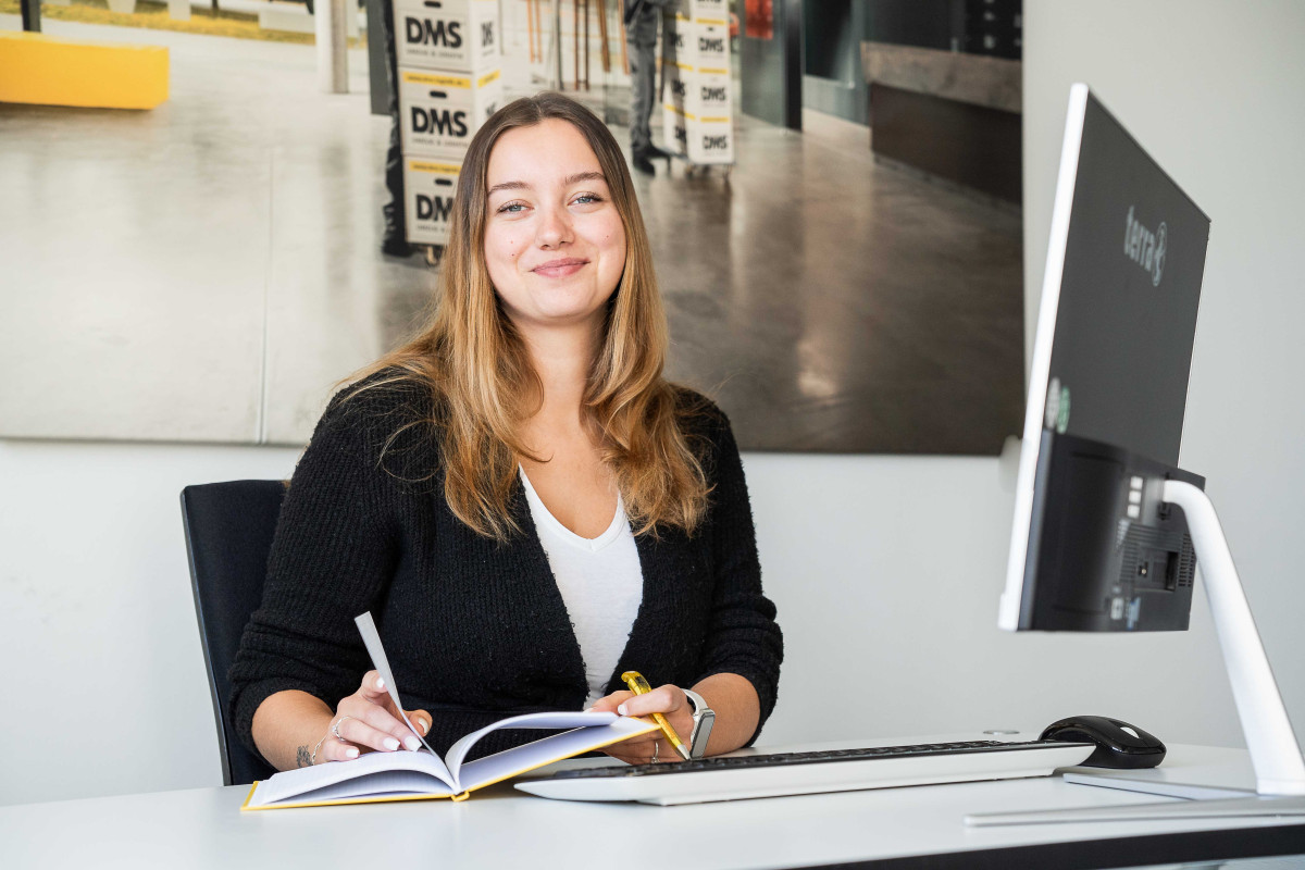 Mitarbeiterin arbeitet an einem Schreibtisch, macht Notizen in einem geöffneten Notizbuch und sitzt vor einer modernen Bürowand mit DMS‑Motiv.