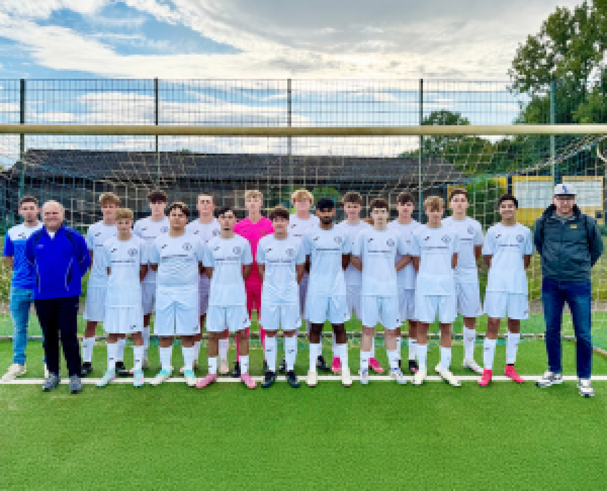 Jugendfußballmannschaft und Trainerteam posieren in weißen Trikots vor dem Tor auf dem Sportplatz.