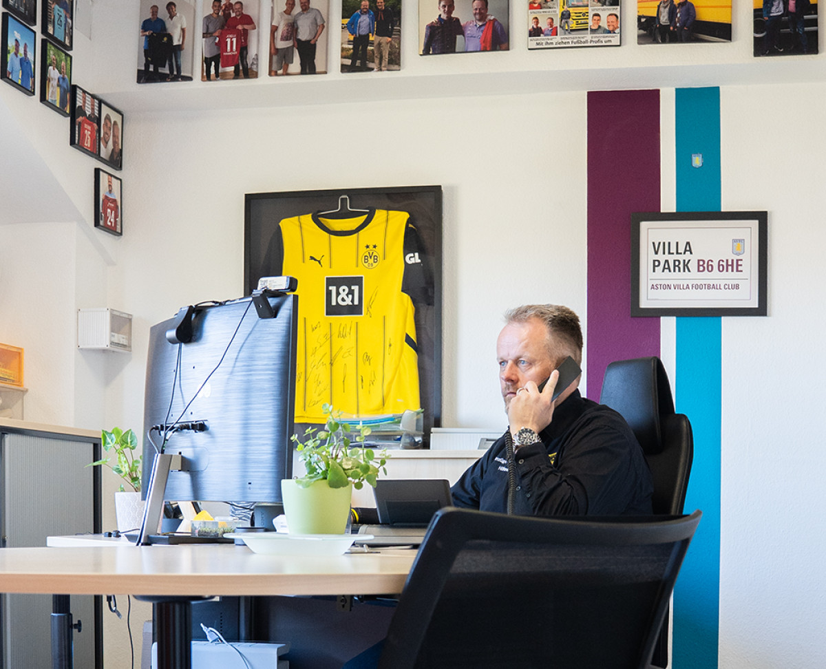 Person führt ein Telefonat am Schreibtisch in einem Büro, dekoriert mit Fußballtrikots und Fanbildern