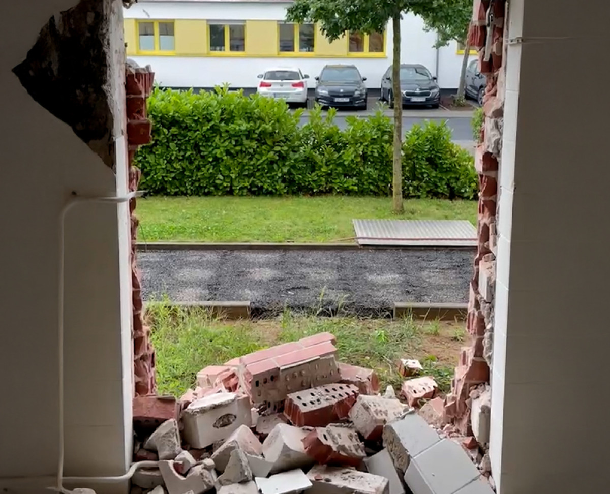 Durchbruch in einer Außenwand mit herausgebrochenen Mauersteinen und Blick auf das Außengelände mit Parkplatz und Büschen