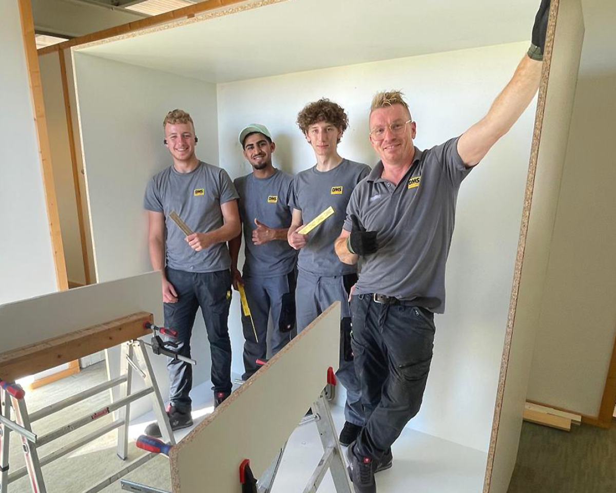 Vier Mitarbeitende in DMS‑Arbeitskleidung montieren Trockenbauwände in einem Innenraum