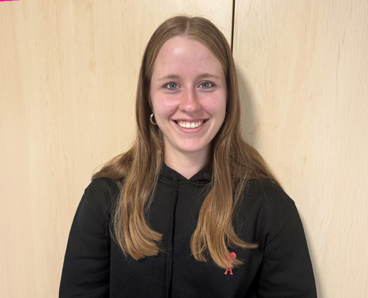 Person mit langen Haaren trägt einen schwarzen Hoodie und steht vor einem Holzschrank.