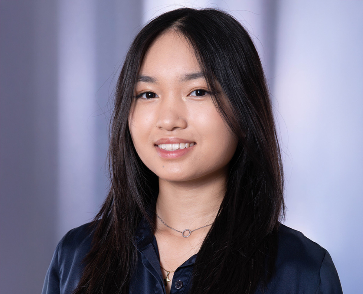 Person mit langen dunklen Haaren trägt eine dunkelblaue Bluse und eine feine Halskette vor einem dezent unscharfen Hintergrund.