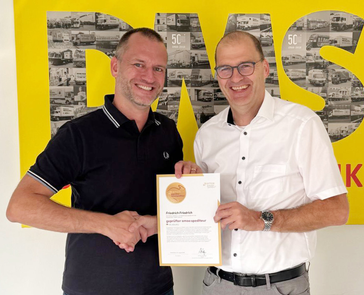 Zwei Personen bei der Übergabe eines Zertifikats vor einem großen gelben DMS‑Hintergrund.