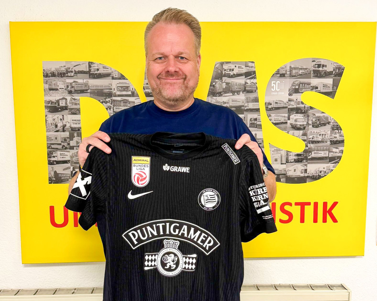 Person hält ein schwarzes Fußballtrikot des SK Sturm Graz vor einem gelben DMS‑Hintergrund.