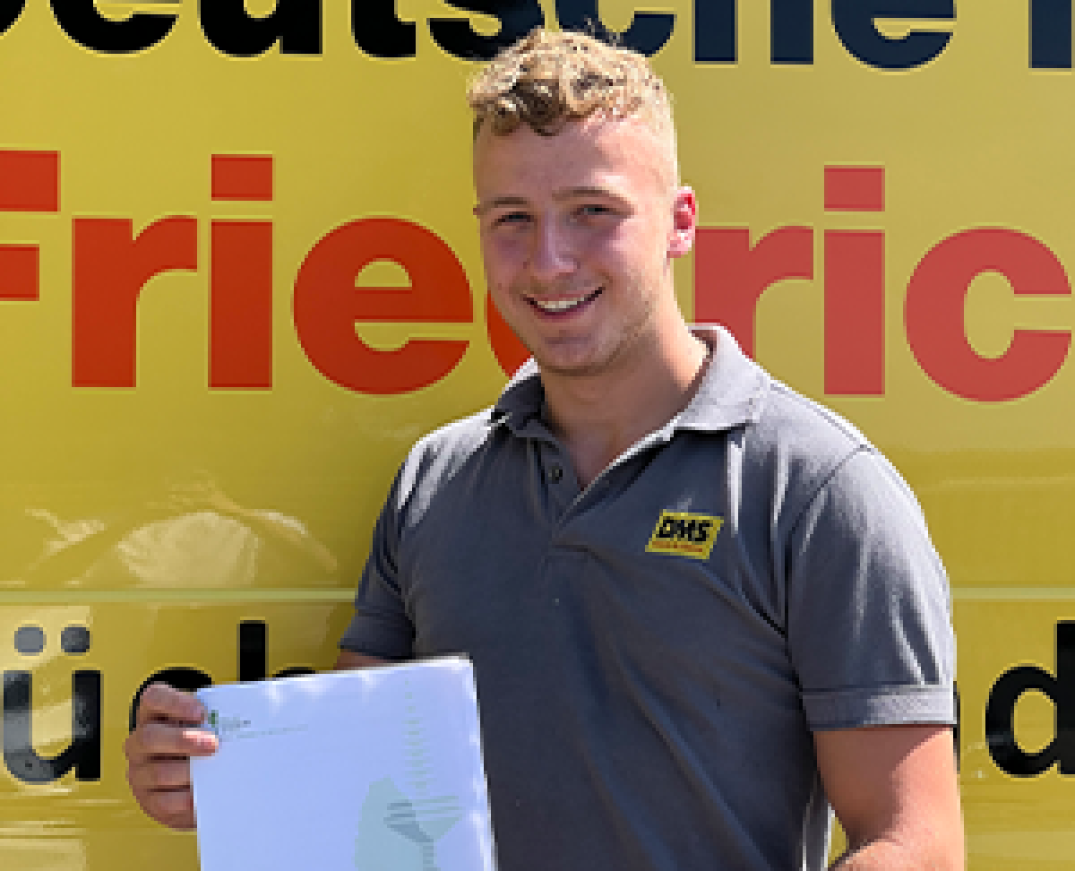 Person in grauem DMS‑Polohemd hält ein Dokument vor einem gelben Firmenfahrzeug von Friedrich Friedrich.