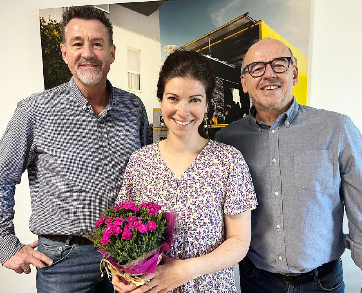 Drei Personen stehen gemeinsam in einem Büro, eine davon hält einen Blumenstrauß.