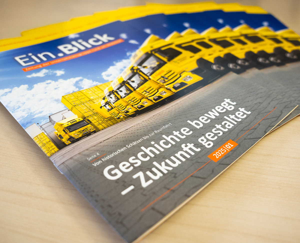 Mehrere Ausgaben des Firmenmagazins ‚EinBlick‘ mit Titelmotiv gelber Lkw‑Flotte und der Überschrift ‚Geschichte bewegt – Zukunft gestaltet‘.
