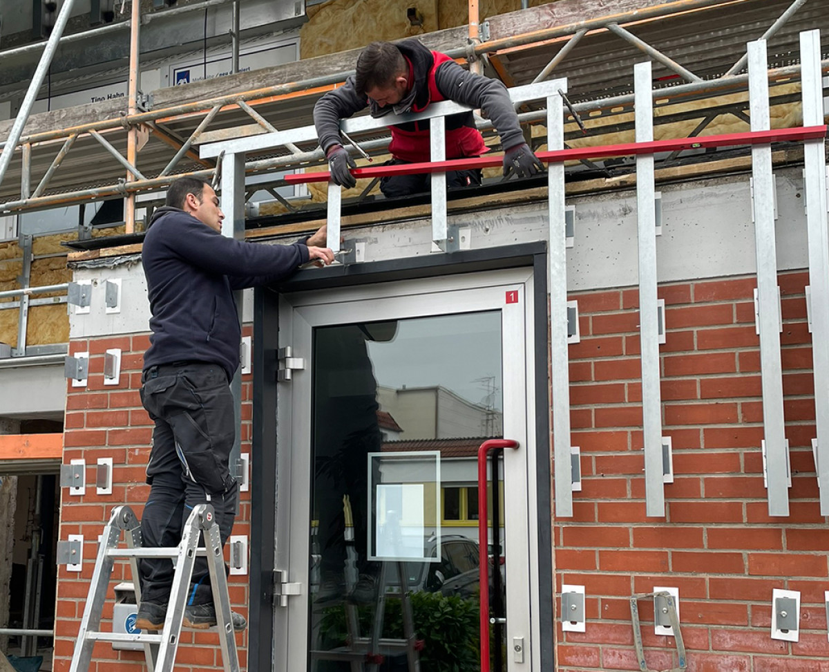 Zwei Handwerker montieren eine Fassadenunterkonstruktion über einem Eingang und arbeiten mit Leiter und Gerüst