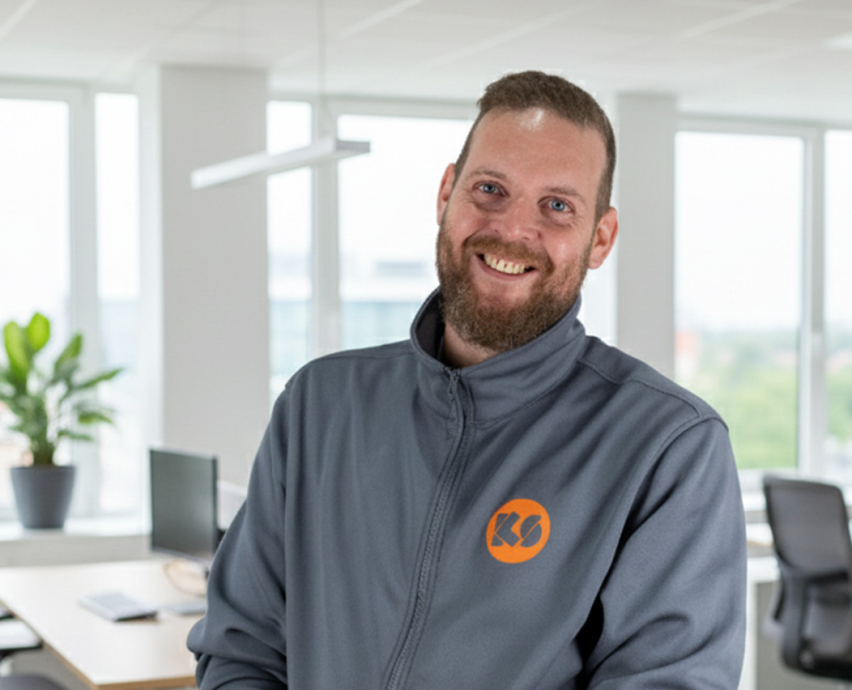 Mitarbeiter in KS‑Arbeitsjacke steht in einem modernen Büro mit großen Fenstern