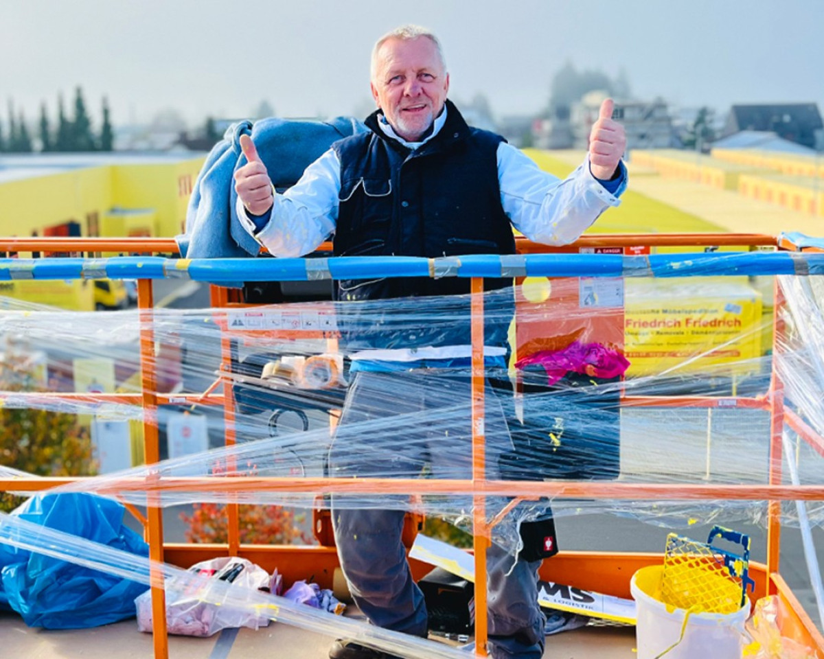 Mitarbeiter zeigt Daumen hoch auf einer Arbeitsbühne vor dem Gelände von Friedrich Friedrich