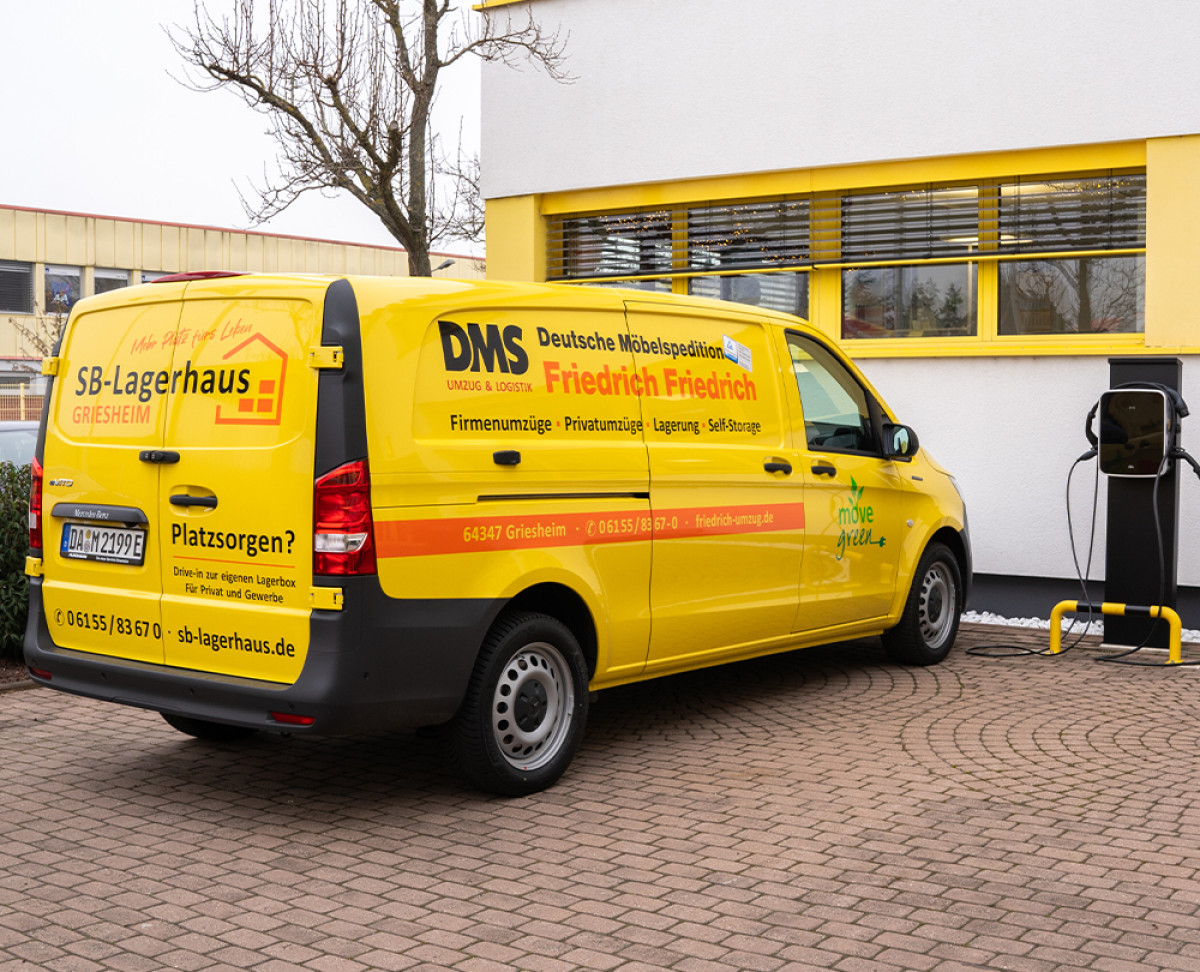 Gelber Elektro‑Transporter der DMS Friedrich Friedrich beim Laden an einer E‑Ladestation vor dem Firmengebäude.