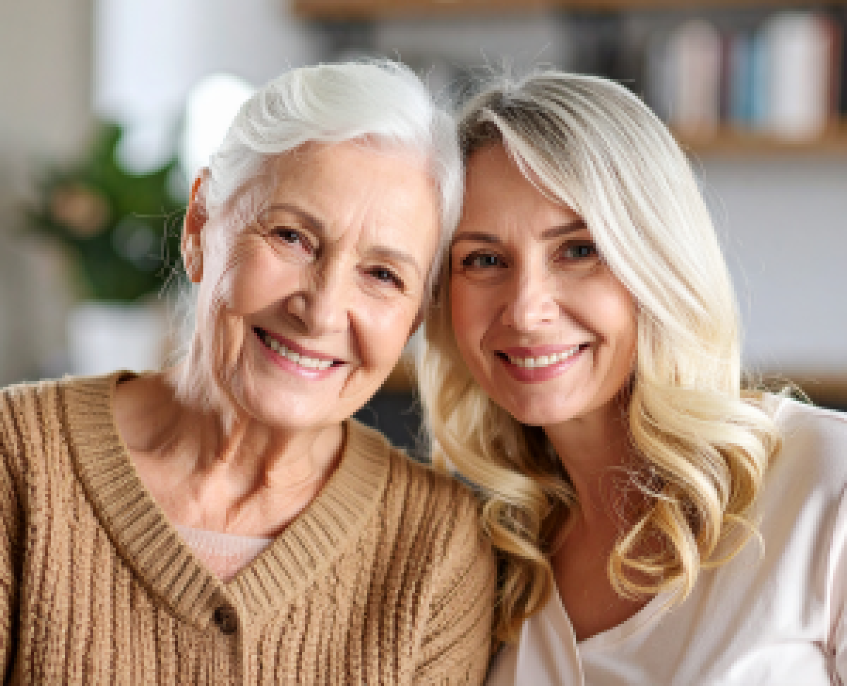 Ältere Frau und jüngere Frau sitzen eng beieinander und lächeln in einem gemütlichen Wohnumfeld
