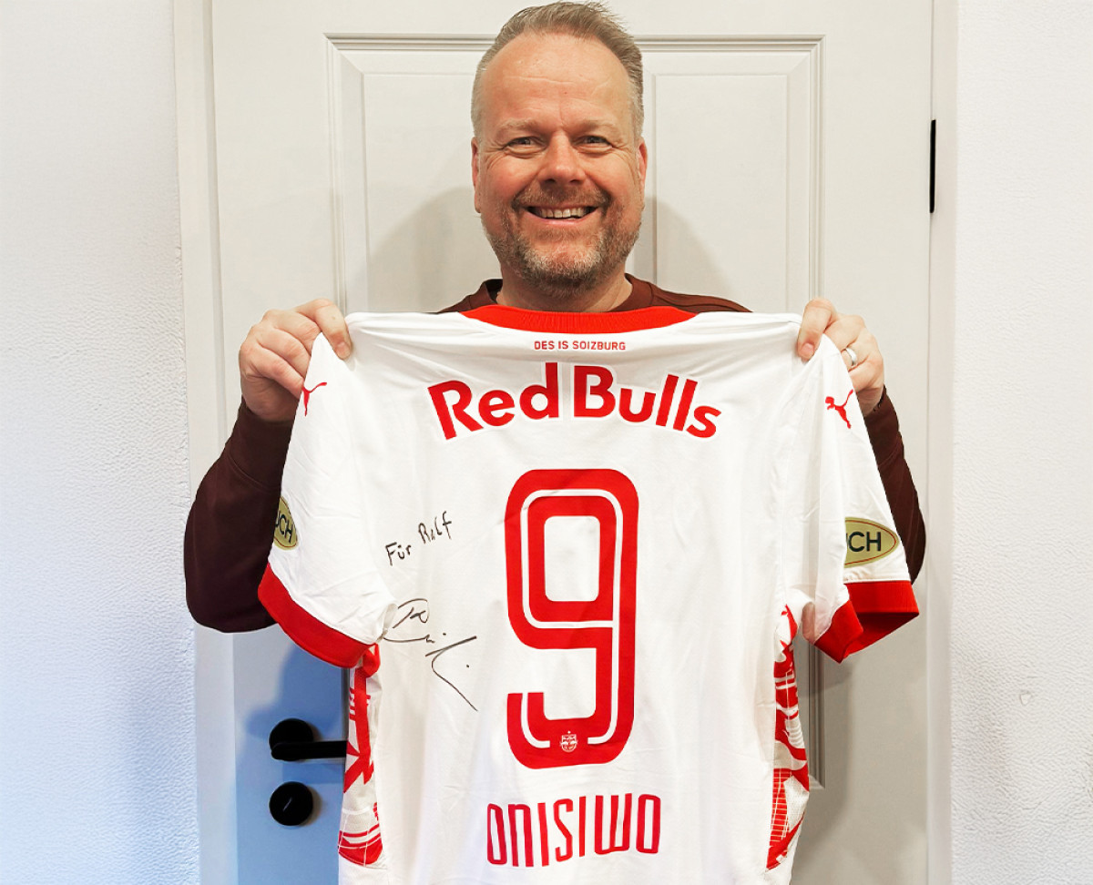 Person hält ein signiertes Red-Bulls-Fußballtrikot mit der Nummer 9 vor einer weißen Tür.