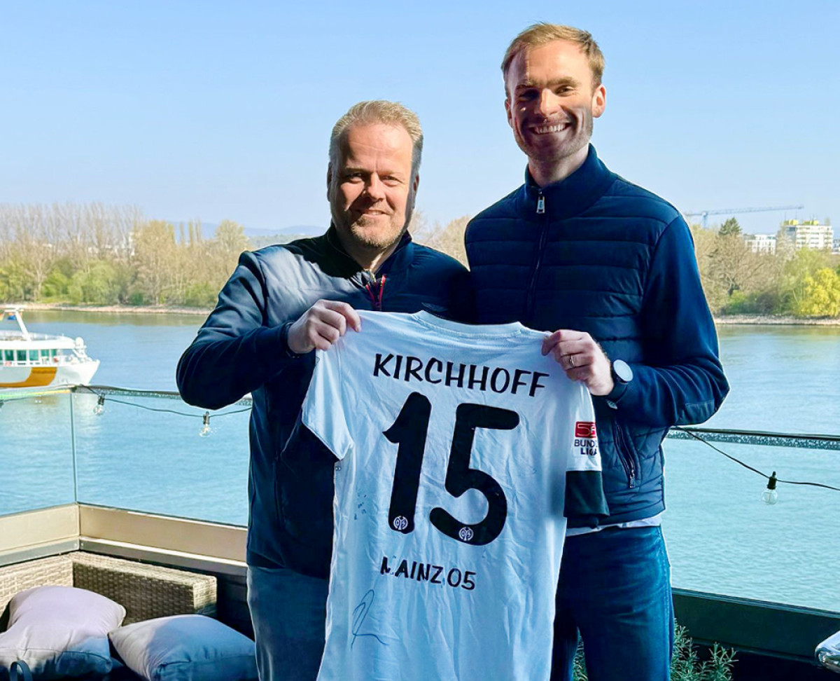 Zwei Personen halten gemeinsam ein weißes Fußballtrikot von Mainz 05 mit der Nummer 15 und dem Namen Kirchhoff vor einer Uferkulisse.   