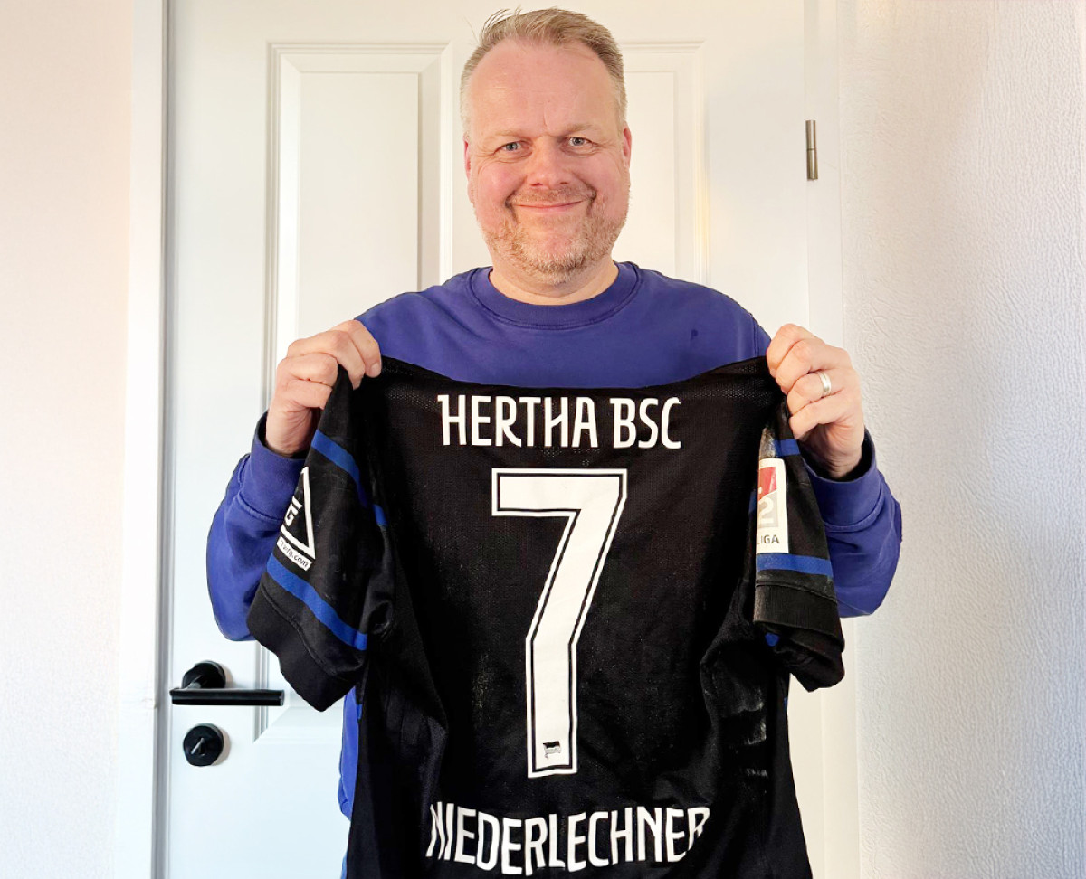 Person hält ein Hertha‑BSC‑Trikot mit der Nummer 7 und dem Namen Niederlechner vor einer weißen Tür.