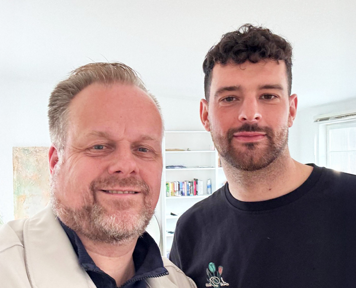 Zwei Personen stehen nebeneinander in einem hellen Raum mit Bücherregal im Hintergrund