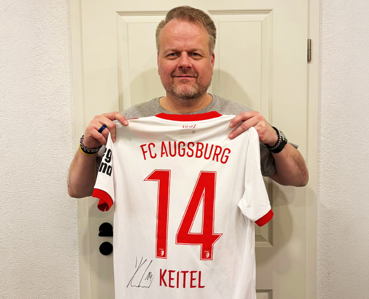Person hält ein signiertes FC‑Augsburg‑Trikot mit der Rückennummer 14 und dem Namen Keitel vor einer Tür.