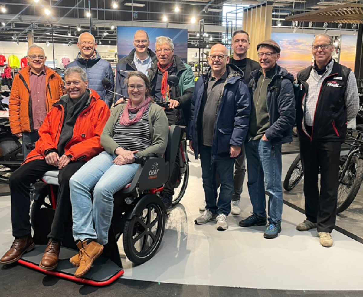 Gruppe von Personen steht in einem Fahrradgeschäft um eine moderne Rollstuhl-Rikscha, in der zwei Personen sitzen.