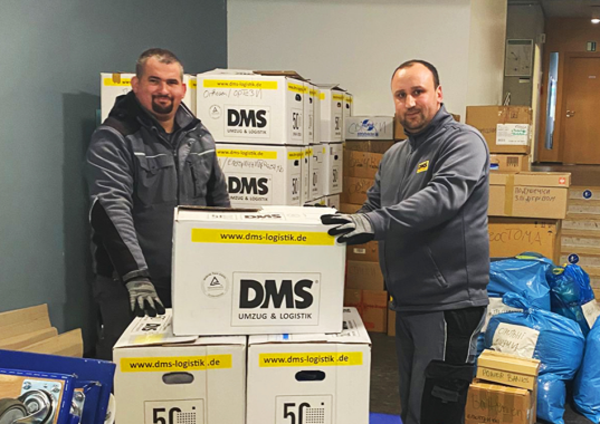 Zwei Mitarbeitende stapeln DMS‑Umzugskartons in einem Raum voller gepackter Hilfsgüter und blauer Säcke.