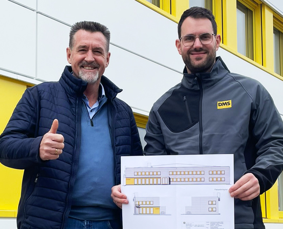 Zwei Personen stehen vor einem Firmengebäude und halten die Baupläne der neuen Fassadengestaltung von Friedrich Friedrich.