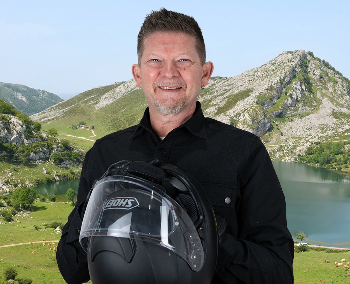 Person hält einen Motorradhelm und steht vor einer Berglandschaft mit See und Felsformationen.