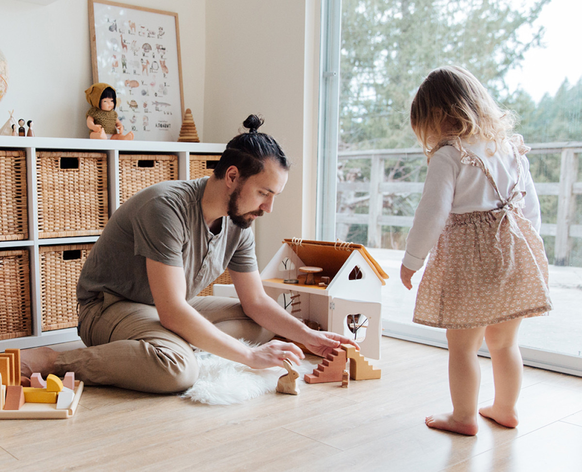 Zwei Personen spielen in einem hellen Wohnzimmer mit einem Holzspielhaus und Bauklötzen auf dem Boden.