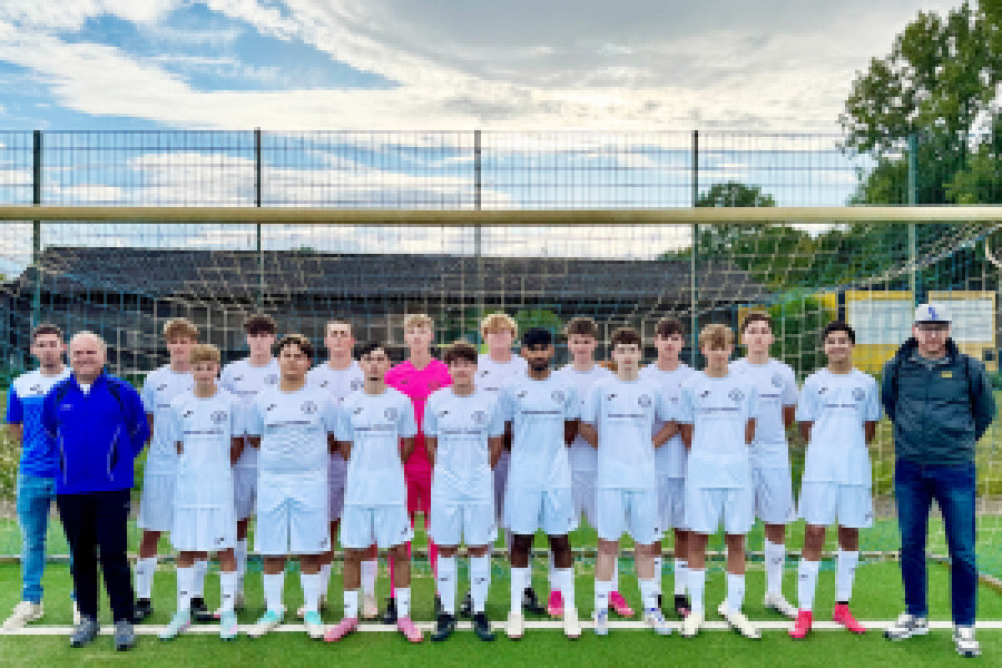 Jugendfußballmannschaft und Trainerteam posieren in weißen Trikots vor dem Tor auf dem Sportplatz.