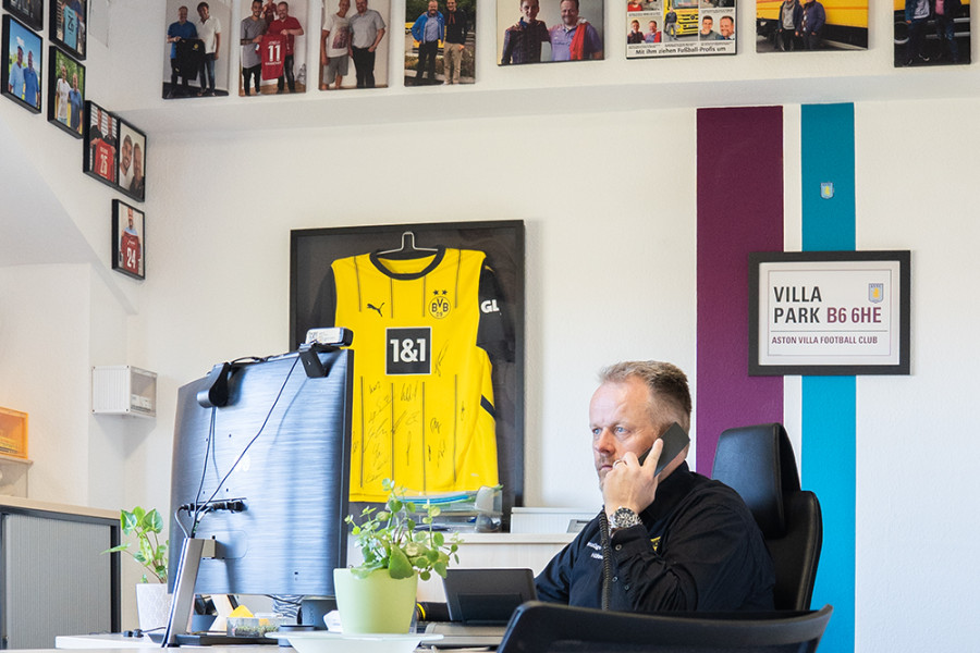 Person führt ein Telefonat am Schreibtisch in einem Büro, dekoriert mit Fußballtrikots und Fanbildern
