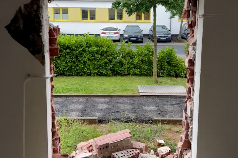 Durchbruch in einer Außenwand mit herausgebrochenen Mauersteinen und Blick auf das Außengelände mit Parkplatz und Büschen