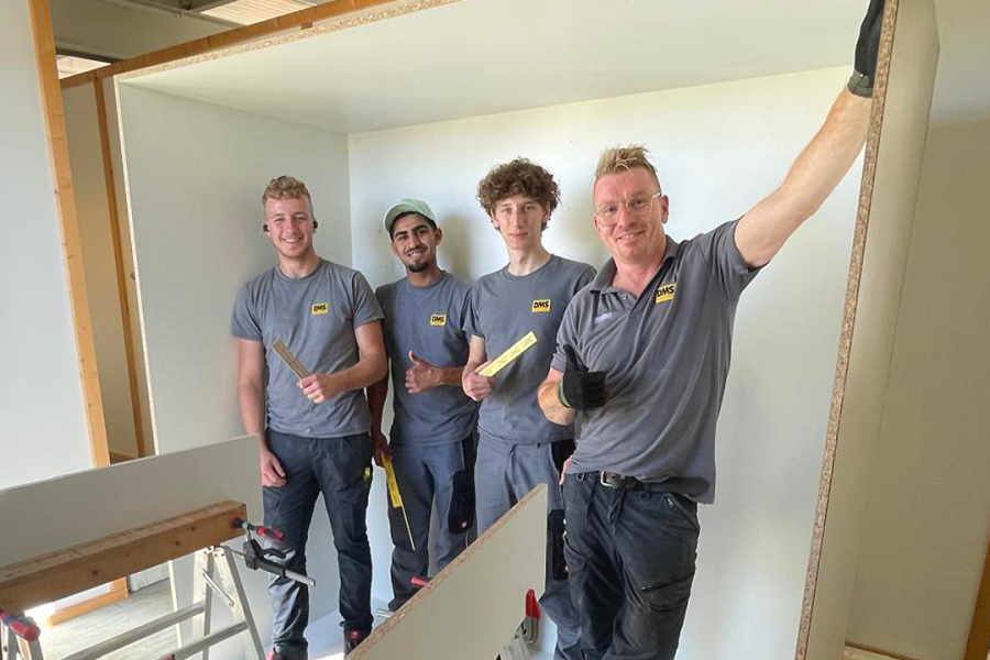 Vier Mitarbeitende in DMS‑Arbeitskleidung montieren Trockenbauwände in einem Innenraum