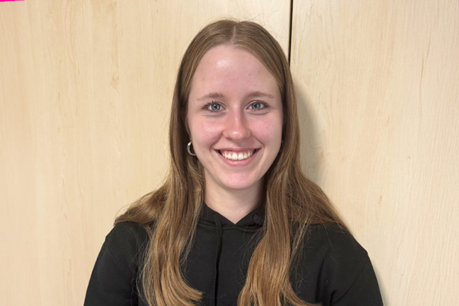 Person mit langen Haaren trägt einen schwarzen Hoodie und steht vor einem Holzschrank.