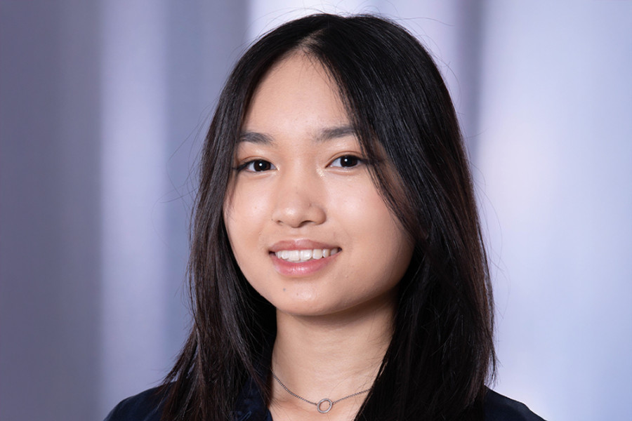 Person mit langen dunklen Haaren trägt eine dunkelblaue Bluse und eine feine Halskette vor einem dezent unscharfen Hintergrund.