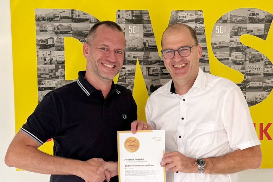 Zwei Personen bei der Übergabe eines Zertifikats vor einem großen gelben DMS‑Hintergrund.