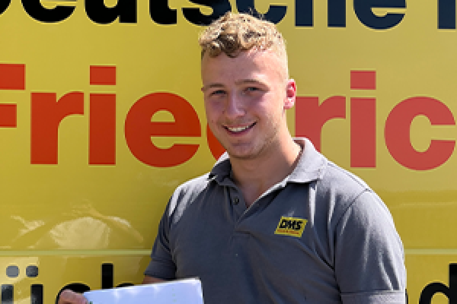 Person in grauem DMS‑Polohemd hält ein Dokument vor einem gelben Firmenfahrzeug von Friedrich Friedrich.