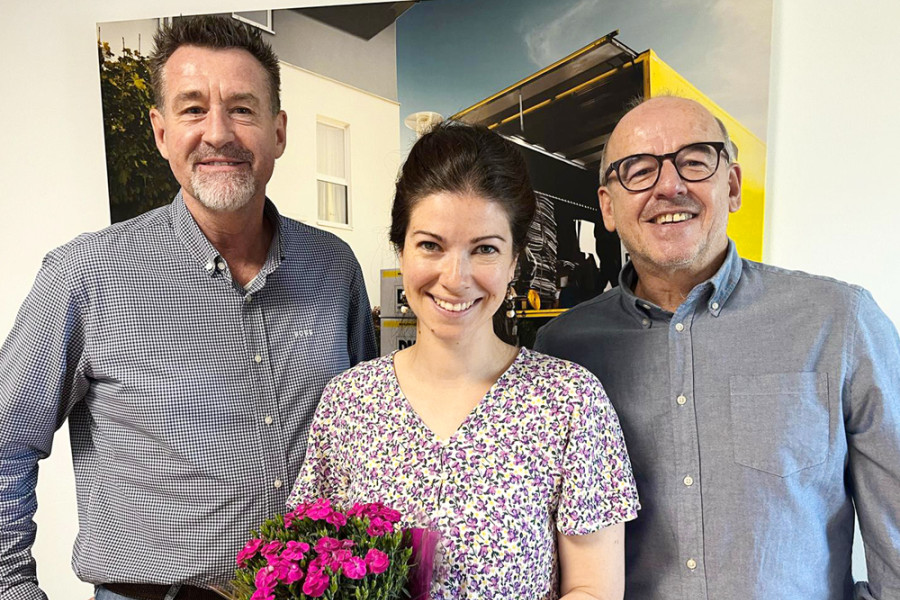 Drei Personen stehen gemeinsam in einem Büro, eine davon hält einen Blumenstrauß.