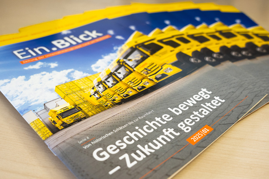Mehrere Ausgaben des Firmenmagazins ‚EinBlick‘ mit Titelmotiv gelber Lkw‑Flotte und der Überschrift ‚Geschichte bewegt – Zukunft gestaltet‘.