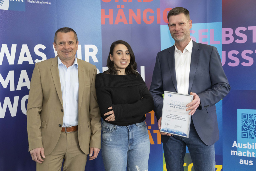 Drei Personen bei einer Auszeichnung, eine Frau in der Mitte, rechts ein Mann mit Urkunde, im Hintergrund eine bunte Wand mit Schrift.