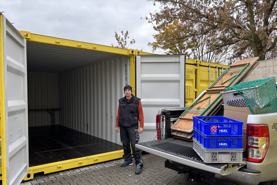 Person steht vor einem geöffneten gelben Lagercontainer, während ein Pickup mit geladenen Transportmaterialien daneben steht.