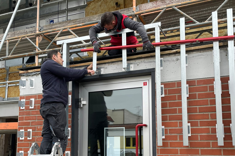 Zwei Handwerker montieren eine Fassadenunterkonstruktion über einem Eingang und arbeiten mit Leiter und Gerüst