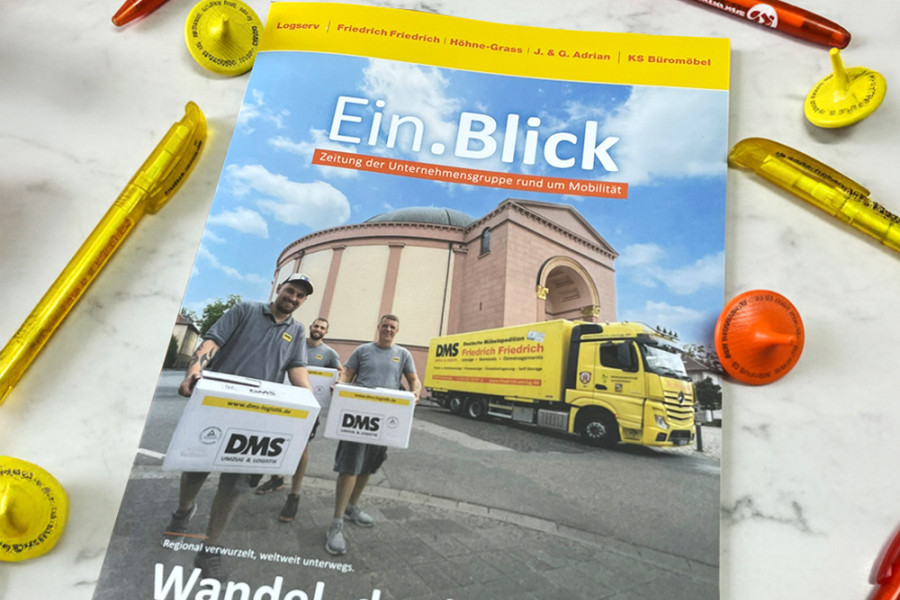 Titelseite der Unternehmenszeitung ‚Ein.Blick‘ mit DMS‑Umzugsteam vor dem Darmstädter Hochzeitsturm und einem gelben Umzugsfahrzeug