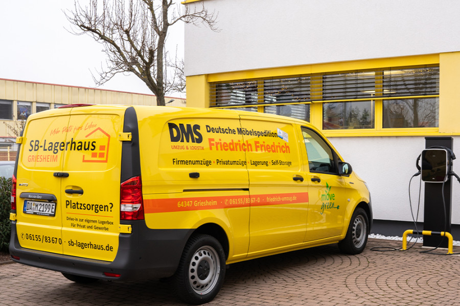 Gelber Elektro‑Transporter der DMS Friedrich Friedrich beim Laden an einer E‑Ladestation vor dem Firmengebäude.