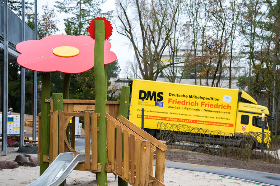 Holzspielanlage auf einem Kita‑Spielplatz mit bunter Blume und einer DMS Friedrich Friedrich Umzugslastwagen im Hintergrund.